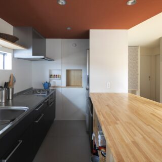 すっきりとしたデザインながら、収納もたっぷりある広いキッチン。天井は赤い珪藻土クロス、床はフレキシブルボードを用い、LDK全体のテイストを揃えた。写真奥にある木製引き戸の小さな窓は玄関につながっている。買い物帰りに食材を手渡しするなど、便利に使える