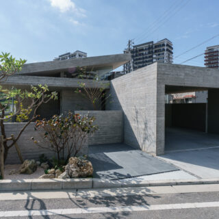 母屋側（東）の外観。右側に車庫、植栽の奥に玄関がある。植物は沖縄由来のものを選び、木々の美しさを堪能できるようアプローチは長めに計画した。コンクリートと緑は相性がいい、と仲本さん。RC造ゆえのコンクリートの外壁が持つ強い印象が、植栽によって和らげられた