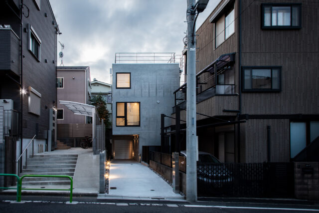 外観。画像中央奥が「武蔵関の住まい」。旗竿地のため道路まで距離があるという条件を生かし、道路に向かって大きく開口した。2階はLDKの窓。3階の窓は法的に必要な非常口でもある。大きさの違う正方形の窓をアクセントに、コンクリートの質感が引き立つデザインの外観とした
