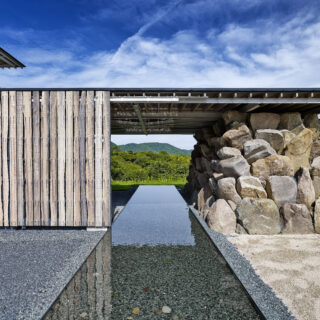 玄関。画像左の木材の壁は玄関の引き戸。水盤を建物に対して直交するように計画、まっすぐ伸びる姿は島まで続く橋のようにも感じられる。画像右は既存の岩を崩れ積みという手法で構造としたもの。屋根のように見える2階のテラスを支えている