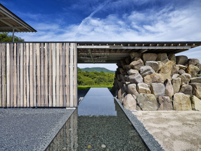 玄関。画像左の木材の壁は玄関の引き戸。水盤を建物に対して直交するように計画、まっすぐ伸びる姿は島まで続く橋のようにも感じられる。画像右は既存の岩を崩れ積みという手法で構造としたもの。屋根のように見える2階のテラスを支えている