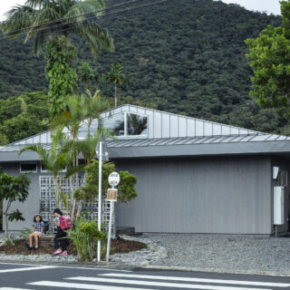 奄美の自然豊かな住宅地に佇むN邸。杉板と入母屋・切妻ハイブリッド屋根の平屋は、周囲の新旧の家々とも馴染んでいる。敷地の一部をバス停のベンチとして開放し、集落とのつながりももたせた。