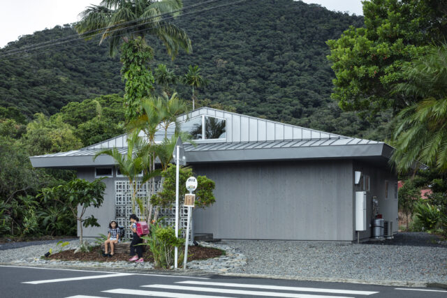 奄美の自然豊かな住宅地に佇むN邸。杉板と入母屋・切妻ハイブリッド屋根の平屋は、周囲の新旧の家々とも馴染んでいる。敷地の一部をバス停のベンチとして開放し、集落とのつながりももたせた。