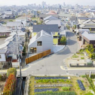 三角形の変形敷地に建物をそっと配置し、空いたスペースを北の駐車場（写真右）や東の庭（写真手前）に活用。マットな明るいシルバーの外壁はガルバリウム鋼板。ウッドフェンスが木の温かみを添え、周囲の長閑な景色に馴染む