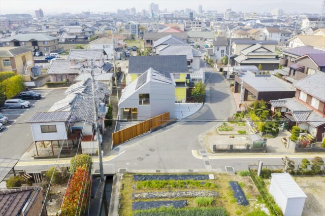 三角形の変形敷地に建物をそっと配置し、空いたスペースを北の駐車場（写真右）や東の庭（写真手前）に活用。マットな明るいシルバーの外壁はガルバリウム鋼板。ウッドフェンスが木の温かみを添え、周囲の長閑な景色に馴染む