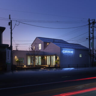 外観夕景（幹線道路側）。アウターデッキの照明と外壁、サインが差別化要素となり、夜間でも目を引くプランだ。奥の2階建てと手前の1階建ての間に、実はデッキが隠れている。外からの視線を遮り、音を遮断する効果も