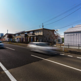 幹線道路側の外観。他社は敷地いっぱいに建てており、外壁も暖色のものが多い。隣地との間に空地（アウターリビング）を設けることで、１団となっている他社との切り分けをおこなっている。断熱性の確保と防音のため、道路側は開口部を最小限にした現実的なプランだ
