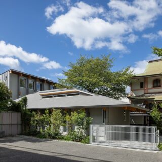 親世帯の外観。ライトグレーと木を合わせたモダンなデザインながら、軒の深い大きな屋根が落ち着いた雰囲気も漂わせている。写真右奥のお寺とも調和する、バランスの良い佇まい