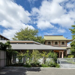 親世帯（写真手前）はお寺（写真奥）と近いため、和の建築とのバランスを考慮して外観をデザイン。外壁はモダンな色使いだが、ゆったりとした大きな屋根と深い軒がお寺の軒と調和し、2つの建物が見事に馴染んでいる
