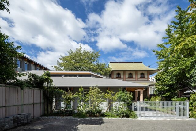 親世帯（写真手前）はお寺（写真奥）と近いため、和の建築とのバランスを考慮して外観をデザイン。外壁はモダンな色使いだが、ゆったりとした大きな屋根と深い軒がお寺の軒と調和し、2つの建物が見事に馴染んでいる