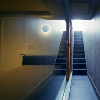 １階、外部通路。画像左、窓は西側中央の居室のもの。奥の階段は２階のバルコニーへ続く。天窓やバルコニーからの光が感じられ、優しい雰囲気。風も抜けるおかげで、プライバシーを守りながら外部が感じられる。階段が室内と外部通路の２か所にあることで、生活の幅が広がった