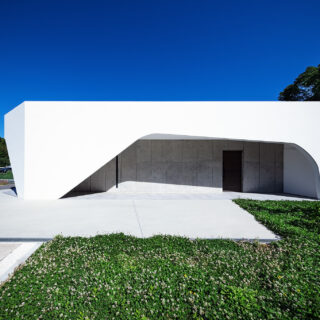 外観。建物手前のコンクリートエリアは駐車場。家の右側に設けたエントランスがある面は白いシェルが庇のように深く広がり、期待感を高める長いアプローチとして計画。壁があることによって、中の躯体を直射や雨汚れから守っている。「機能とデザインがリンクする」と石さん