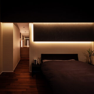 主寝室。床や壁に暗目の色を選び、夜間は高級感あるしっとりした雰囲気を楽しめるようにした。奥へ進むとシューズクローゼットやウォークインクローゼットに直接アクセスできる。背面は広々とした庭があり、LDKまで見通せる。自分の家を眺めくつろぐひとときは贅沢極まりない