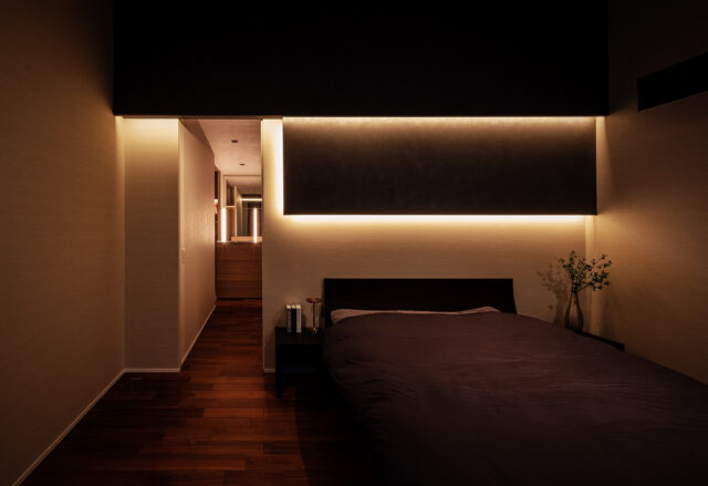 主寝室。床や壁に暗目の色を選び、夜間は高級感あるしっとりした雰囲気を楽しめるようにした。奥へ進むとシューズクローゼットやウォークインクローゼットに直接アクセスできる。背面は広々とした庭があり、LDKまで見通せる。自分の家を眺めくつろぐひとときは贅沢極まりない