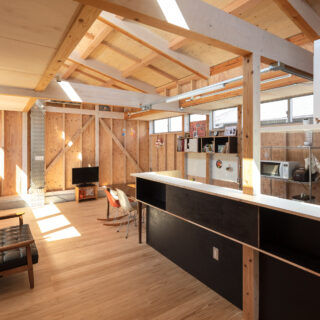 LDK。外観からは想像できないほど明るく開放的な室内。画像左の低い天井から壁を立ち上げ、ハイサイドライトを計画。豊かに日射が入る。木材の中に配管などの金属をポイント使いした設えは、日々の生活で自然に生まれた雑多な雰囲気を大らかに受け止める