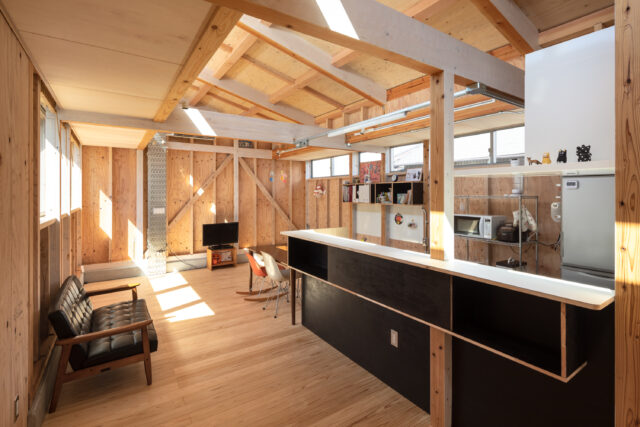 LDK。外観からは想像できないほど明るく開放的な室内。画像左の低い天井から壁を立ち上げ、ハイサイドライトを計画。豊かに日射が入る。木材の中に配管などの金属をポイント使いした設えは、日々の生活で自然に生まれた雑多な雰囲気を大らかに受け止める