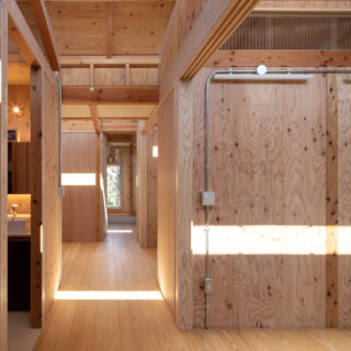 玄関から室内を見る。3つのハイサイドライトからの光がリズミカルに落ち、奥へ奥へと意識をいざなう。最奥に窓を設けたおかげで、より奥行きが感じられるようになった。柱や梁、合板耐力壁など構造体表しの室内。シルバーの無機質な配管が空間の印象に変化を与える