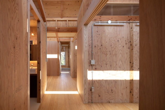 玄関から室内を見る。3つのハイサイドライトからの光がリズミカルに落ち、奥へ奥へと意識をいざなう。最奥に窓を設けたおかげで、より奥行きが感じられるようになった。柱や梁、合板耐力壁など構造体表しの室内。シルバーの無機質な配管が空間の印象に変化を与える