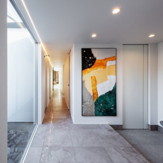 ゲストルーム前からLDK方向を見る。右の仕切り戸はシューズクローゼットのもの。シューズクローゼットからは廊下以外に、主寝室やウォークインクローゼットにもアクセスできる。廊下とLDKを仕切る扉はガラス戸としたうえ、天井右側にラインで走る照明を取り入れ奥行きを強調