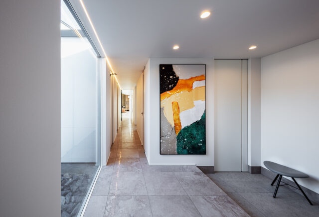 ゲストルーム前からLDK方向を見る。右の仕切り戸はシューズクローゼットのもの。シューズクローゼットからは廊下以外に、主寝室やウォークインクローゼットにもアクセスできる。廊下とLDKを仕切る扉はガラス戸としたうえ、天井右側にラインで走る照明を取り入れ奥行きを強調