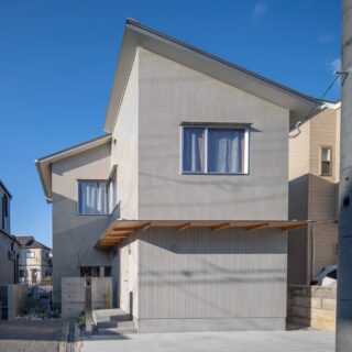 前方に片流れ屋根、奥に切妻屋根をもつL字型のS邸。グレーのモルタルで仕上げられた静謐な佇まい。大きな庇の下の壁面には、耐光塗装を施した杉板がアクセントを添えている。
