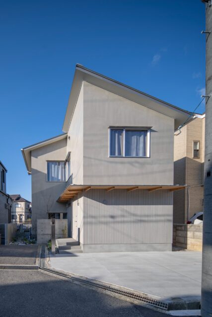 前方に片流れ屋根、奥に切妻屋根をもつL字型のS邸。グレーのモルタルで仕上げられた静謐な佇まい。大きな庇の下の壁面には、耐光塗装を施した杉板がアクセントを添えている。