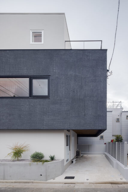 リビングのある２階部分を強調するため、白い箱にグレーの箱が一部重なるような構成とした。これにより、駐車スペースに軒がかかり、雨に濡れずに乗り降りできるように。