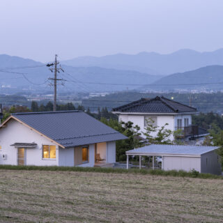 遠くの空に山のシルエットが浮かぶ夕景の中、温かな光を灯す「西春近の家」。西から見ると切妻のきれいなラインがいっそう引き立つ