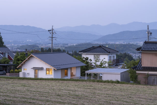 遠くの空に山のシルエットが浮かぶ夕景の中、温かな光を灯す「西春近の家」。西から見ると切妻のきれいなラインがいっそう引き立つ