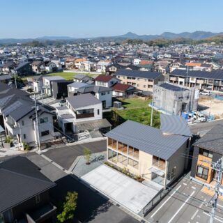 上空から見た建物（右から２番目）。市道に面する南側に駐車場、他にも敷地内に中庭など２つの余白を設け、その余白を躱すように建物を「くの字」に配置した。建物の厚みを薄くし、近隣の家々と同程度の印象を受けるようなボリュームに抑えて地域に調和させた