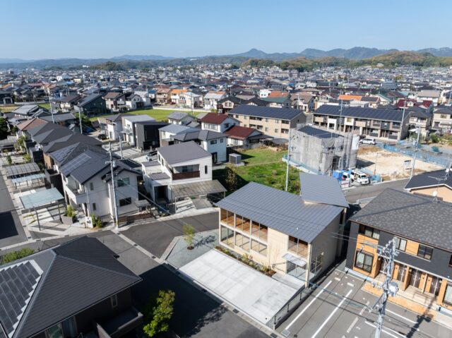 上空から見た建物（右から２番目）。市道に面する南側に駐車場、他にも敷地内に中庭など２つの余白を設け、その余白を躱すように建物を「くの字」に配置した。建物の厚みを薄くし、近隣の家々と同程度の印象を受けるようなボリュームに抑えて地域に調和させた