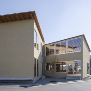 北西から見た外観。左端の木を残したいという要望から、北側に庭を計画。さらに西側に中庭も設けた。くの字の建物のうち、右側１階が機能訓練室。掃き出し窓からアクセスし、中庭をより豊かに活用できるように設えた。利用者様スタッフさんみんなで庭づくりを楽しまれている