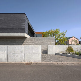 北西から見た外観。外壁には２階部分はサイディング、1階部分はコンクリートを採用した。コンクリートは杉板柄など３種類が使い分けられ表情豊か。外構のコンクリート壁を一部下げ、壁の前に植栽帯を計画（画像右）し、壁の印象をやわらげた。壁の間を入ると家族用の駐車場に進む