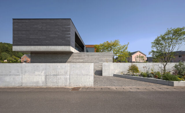 北西から見た外観。外壁には２階部分はサイディング、1階部分はコンクリートを採用した。コンクリートは杉板柄など３種類が使い分けられ表情豊か。外構のコンクリート壁を一部下げ、壁の前に植栽帯を計画（画像右）し、壁の印象をやわらげた。壁の間を入ると家族用の駐車場に進む