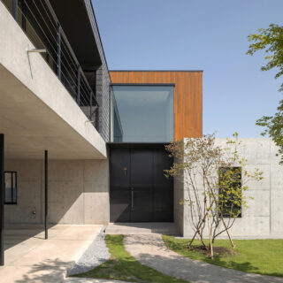 リビング前の庭、アプローチから玄関を見る。奥さまのご希望により玄関は約3.2ｍの鉄製のものを採用。玄関上にあるのが吹き抜けの開口。屋根を薄くするなど工夫し「ガラスの箱がついているように見せたかった」とのこと。画像左は駐車場。雨の日でも濡れずに室内に入れると好評だ