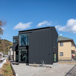 接道部分から見た家。左は川と記念碑、右は公園と変形敷地ながら恵まれた条件の土地といえる。小さな正方形をまず配置し、余白に水回り（左の突き出した部分）玄関（手前部分）、庭などを設けた。余白にゆとりがあるおかげで、様々な用途に応用できる