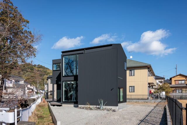 接道部分から見た家。左は川と記念碑、右は公園と変形敷地ながら恵まれた条件の土地といえる。小さな正方形をまず配置し、余白に水回り（左の突き出した部分）玄関（手前部分）、庭などを設けた。余白にゆとりがあるおかげで、様々な用途に応用できる