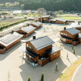 西側高所からの全景。左側に並ぶのはA棟、右に2つ並ぶ棟はB棟。収容人数は同じながら、大きな平屋のA棟に対して、B棟は高さを抑えたコンパクトな2階建て。B棟の2階からは、A棟からも見える美しい山が眺められる。場の持つイメージを踏まえ、宿泊施設は全て切妻屋根とした
