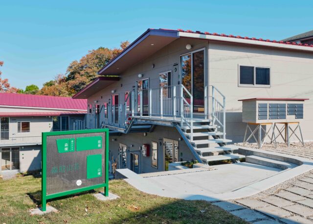 緑に映える赤い屋根、地形を生かした個性的なデザインが目を引く。敷地の入口では、3棟の配置を示す案内板が訪れた人をお出迎え。写真右、建物手前にある郵便受けはウミネコアーキでDIY。イームズチェアを思わせる脚は百葉箱用のもの。センスあふれる素材使いが楽しい