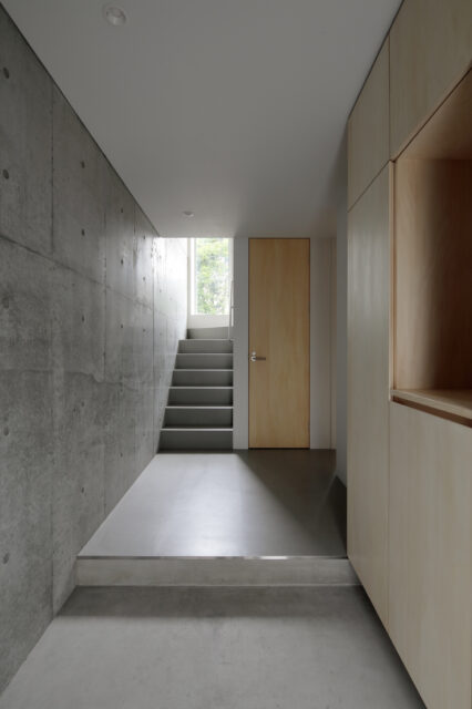 RC造のコンクリート打ち放しの玄関。地階には寝室と洗面室・浴室といった水回りを配置。階段の先にあるのが、庭へ向かう第二の出入り口。ガラス扉にしたことで、庭からの光が降りてくる。