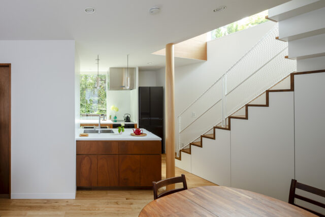 1階ダイニングからキッチン方向を見る。画像左に玄関への扉が見える。キッチンの奥にも玄関への扉を計画し、回遊性を持たせた。奥さまは玄関からキッチンを通り階段へアクセスできる。階段下は収納。介護用品など、散らかりやすいものをさっと仕舞えて便利