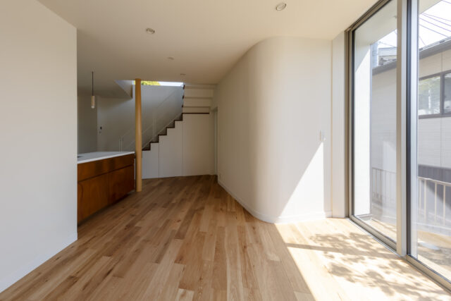 1階。車椅子を使用するため、床は傷に強い塗装を施した素材を採用。巾木もやや高めに設定し、壁の汚れを防ぐ。右の壁面は一部アールに。光が柔らかく室内に広がり、自然と屋外へ視線が伸びる。壁面の裏は水回り。あえて南側に配置し、明るく快適な水回り空間を実現