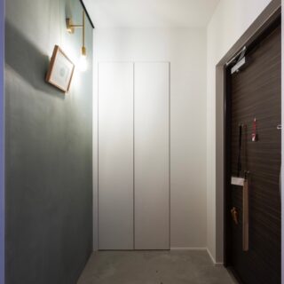 玄関。入口正面はブルーグレーの漆喰壁を背景としたピクチャーレール付きウォールで、真鍮ブラケット照明と共に住宅の第一印象を彩る。