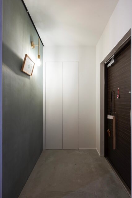 玄関。入口正面はブルーグレーの漆喰壁を背景としたピクチャーレール付きウォールで、真鍮ブラケット照明と共に住宅の第一印象を彩る