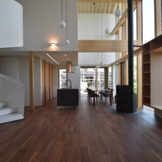 １階LDK。画像左、螺旋階段の手前と奥は玄関に繋がる。回遊性ある動線は移動がしやすい。奥の黒いカウンター部分がキッチン。カウンターの反対側に三枚扉の収納を設け、冷蔵庫もそこに収納。螺旋階段の奥の壁面の扉はパントリー。最奥の窓からは義理のご家族の家が見える