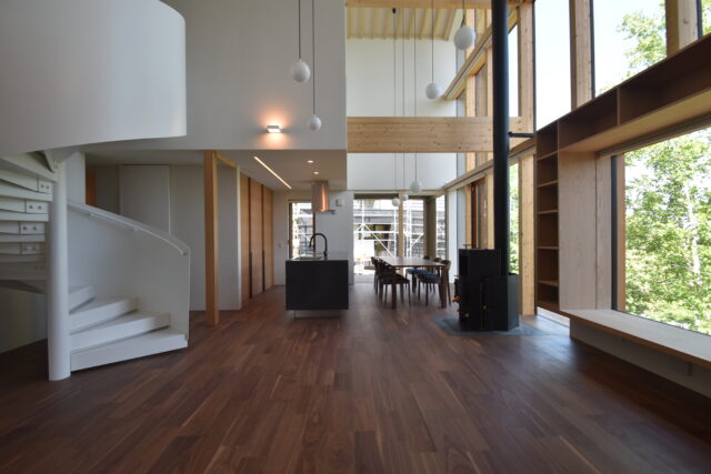 １階LDK。画像左、螺旋階段の手前と奥は玄関に繋がる。回遊性ある動線は移動がしやすい。奥の黒いカウンター部分がキッチン。カウンターの反対側に三枚扉の収納を設け、冷蔵庫もそこに収納。螺旋階段の奥の壁面の扉はパントリー。最奥の窓からは義理のご家族の家が見える