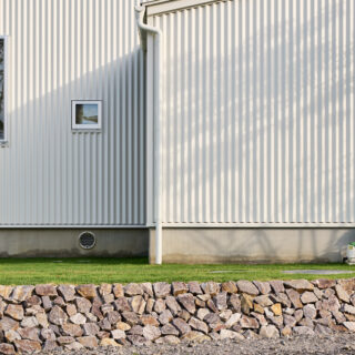 集落側から見た外壁。基礎の丸い穴が、エアサイクルの通気口だ。庭の木の影がガルバリウムの外壁に柔らかに映る