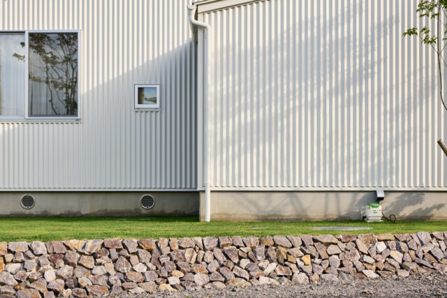 集落側から見た外壁。基礎の丸い穴が、エアサイクルの通気口だ。庭の木の影がガルバリウムの外壁に柔らかに映る
