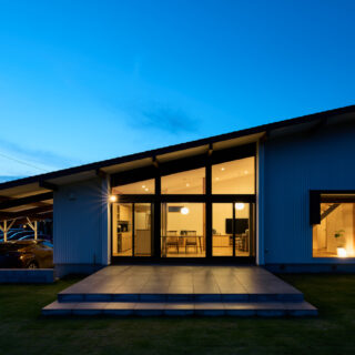 夕景外観。26ｍの長さを持つ片流れの屋根形状がよくわかる。屋根が高い部分のみ、一部2階建て。手前の広い庭には広大なテラスで、仲間と集まりBBQなどを楽しむための場所だ。手前が川で人通りもないため、カーテンなどを閉めずに、常にこの状態で景色を楽しんでいるそうだ