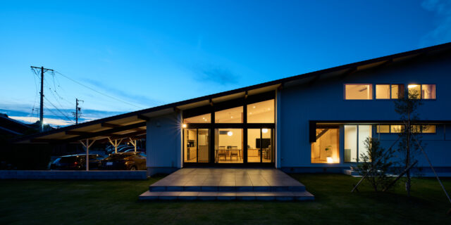 夕景外観。26ｍの長さを持つ片流れの屋根形状がよくわかる。屋根が高い部分のみ、一部2階建て。手前の広い庭には広大なテラスで、仲間と集まりBBQなどを楽しむための場所だ。手前が川で人通りもないため、カーテンなどを閉めずに、常にこの状態で景色を楽しんでいるそうだ